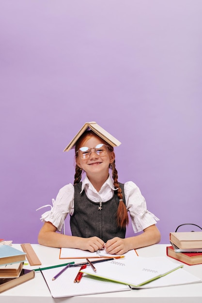 Lächelndes junges Schulmädchen mit Ingwerzöpfen mit Brille und Buch auf dem Kopf in klassischem Outfit, das posiert, während es am Schreibtisch sitzt