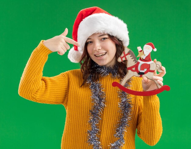 Lächelndes junges slawisches Mädchen mit Weihnachtsmütze und mit Girlande um den Hals, das den Weihnachtsmann auf Schaukelpferddekoration hält und zeigt, isoliert auf grüner Wand mit Kopierraum