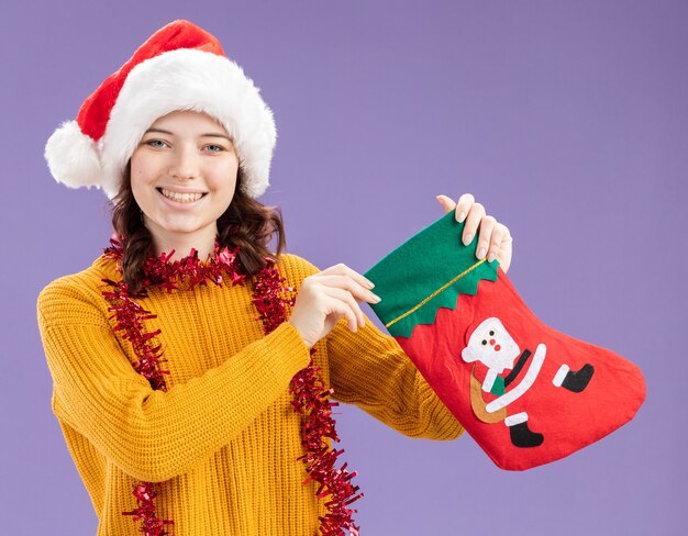 lächelndes junges slawisches Mädchen mit Weihnachtsmütze und mit Girlande um Hals, der Weihnachtsstrumpf lokalisiert auf lila Hintergrund mit Kopienraum hält