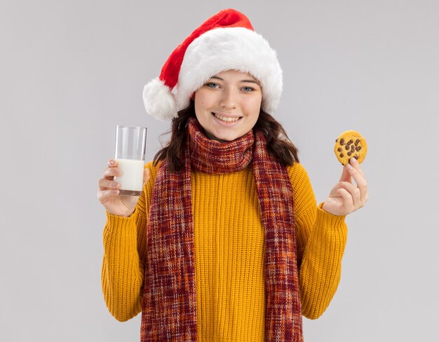 Lächelndes junges slawisches Mädchen mit Weihnachtsmütze und mit Schal um den Hals, das ein Glas Milch und Kekse hält, isoliert auf weißer Wand mit Kopierraum
