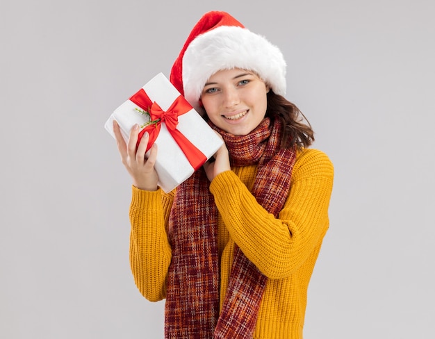 Kostenloses Foto lächelndes junges slawisches mädchen mit weihnachtsmütze und mit schal um hals, der weihnachtsgeschenkbox lokalisiert auf weißem hintergrund mit kopienraum hält