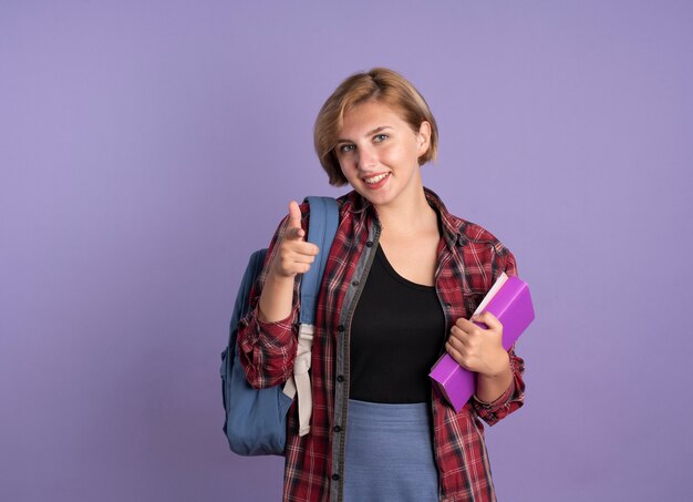 Lächelndes junges slawisches Studentenmädchen, das Rucksack trägt, hält Buch- und Notizbuchpunkte an der Kamera