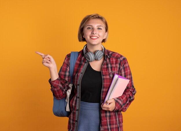 Lächelndes junges slawisches Studentenmädchen mit Kopfhörern, das einen Rucksack trägt, der ein Buch und ein Notizbuch hält, die auf die Seite zeigen