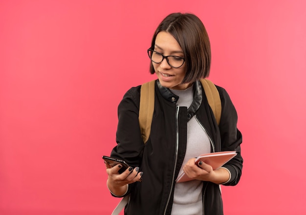 Lächelndes junges Studentenmädchen, das Brille und Rückentasche hält, die Notizblock und Handy betrachten, das Telefon lokalisiert auf rosa Wand betrachtet