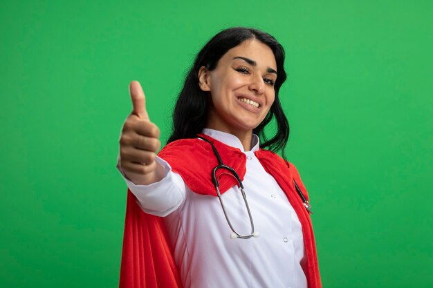 Lächelndes junges Superheldenmädchen, das medizinisches Gewand mit Stethoskop trägt, zeigt Daumen oben lokalisiert auf Grün