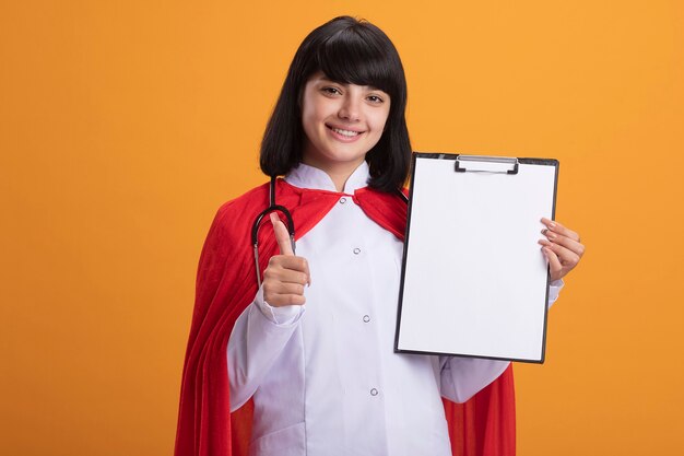 Lächelndes junges Superheldenmädchen, das Stethoskop mit medizinischem Gewand und Umhang hält, das Klemmbrett hält, das Daumen oben auf orange Wand lokalisiert zeigt