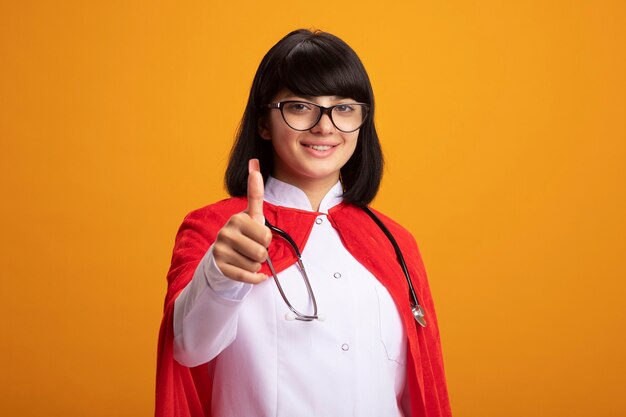 Lächelndes junges Superheldenmädchen, das Stethoskop mit medizinischem Gewand und Umhang mit Brille trägt, zeigt Daumen oben isoliert auf Orange