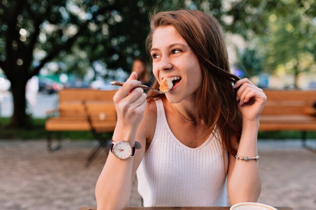 Lächelndes kaukasisches weibliches Modell, das ausgefallenen Cupcake in der Sommercafeteria im Freien isst