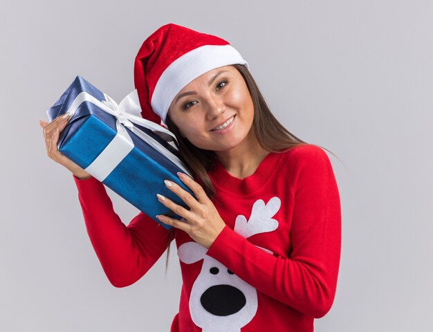 Lächelndes kippendes junges asiatisches Mädchen des Kippkopfes, das Weihnachtshut mit Pullover hält, der Geschenkbox um Gesicht lokalisiert auf weißem Hintergrund hält