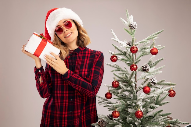 Lächelndes, kippendes junges schönes Mädchen, das in der Nähe des Weihnachtsbaums steht und Weihnachtsmütze mit Brille trägt, die Geschenkbox isoliert auf weißem Hintergrund hält holding