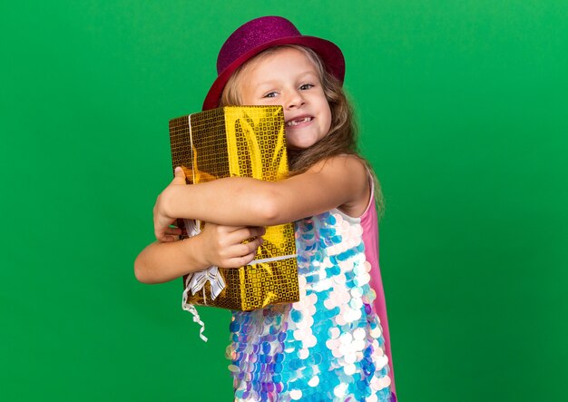 lächelndes kleines blondes Mädchen mit lila Partyhut umarmt Geschenkbox lokalisiert auf grüner Wand mit Kopienraum