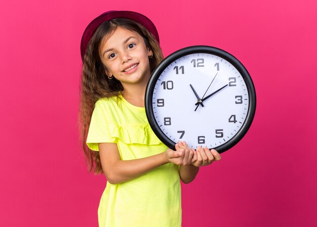 Lächelndes kleines kaukasisches Mädchen mit lila Partyhut, das die Uhr isoliert auf rosa Wand mit Kopienraum hält
