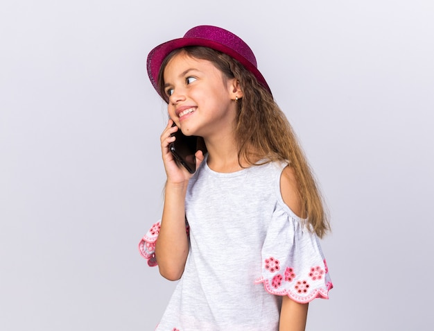 lächelndes kleines kaukasisches Mädchen mit lila Partyhut, das Seite betrachtet, die am Telefon spricht, lokalisiert auf weißer Wand mit Kopienraum