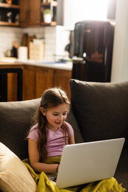 Lächelndes kleines Mädchen, das auf dem Sofa sitzt und Laptop im Wohnzimmer benutzt