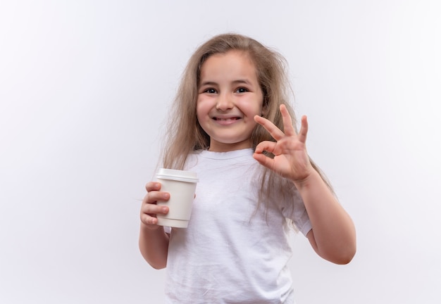 Lächelndes kleines Schulmädchen, das weißes T-Shirt hält, das Tasse Kaffee hält, die okey Geste auf lokalisiertem weißem Hintergrund zeigt