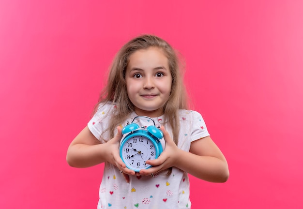 Lächelndes kleines Schulmädchen, das weißes T-Shirt hält, das Wecker auf lokalisiertem rosa Hintergrund hält