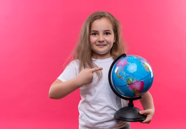 Lächelndes kleines Schulmädchen, das weißes T-Shirt hält Globus auf lokalisiertem rosa Hintergrund trägt