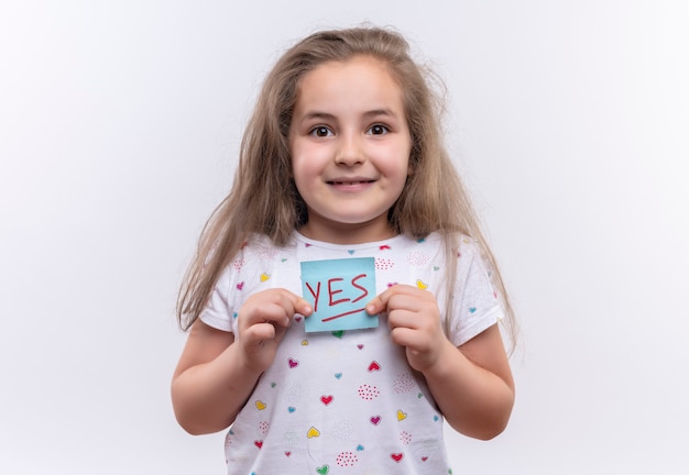 Lächelndes kleines Schulmädchen, das weißes T-Shirt hält Papiermarke auf lokalisiertem weißem Hintergrund hält