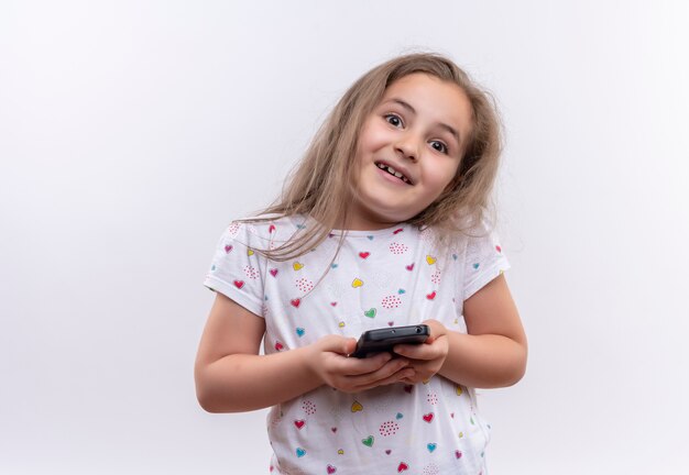 Lächelndes kleines Schulmädchen, das weißes T-Shirt hält Telefon auf lokalisiertem weißem Hintergrund hält