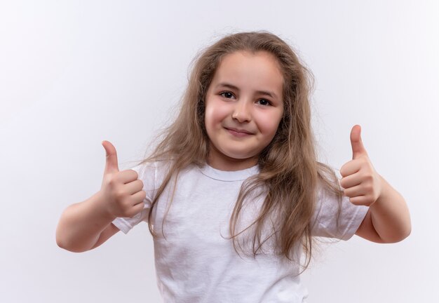 Lächelndes kleines Schulmädchen, das weißes T-Shirt ihre Daumen oben auf lokalisiertem weißem Hintergrund trägt