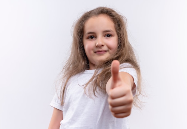 Lächelndes kleines Schulmädchen, das weißes T-Shirt ihren Daumen oben auf lokalisiertem weißem Hintergrund trägt