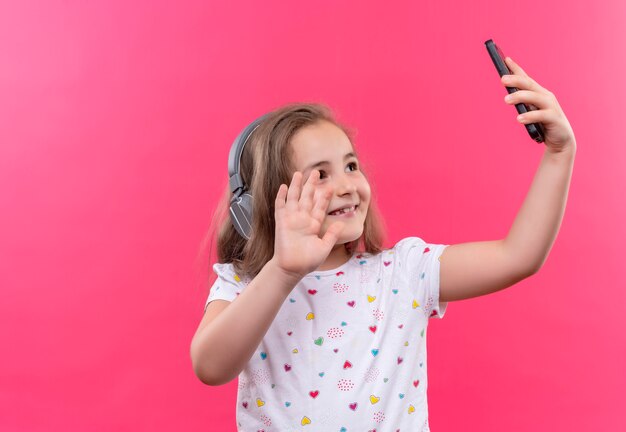 Lächelndes kleines Schulmädchen, das weißes T-Shirt im Kopfhörer trägt, spricht am Telefon auf lokalisiertem rosa Hintergrund