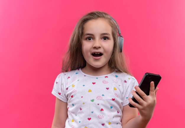 Lächelndes kleines Schulmädchen, das weißes T-Shirt in den Kopfhörern hält Telefon auf lokalisiertem rosa Hintergrund hält