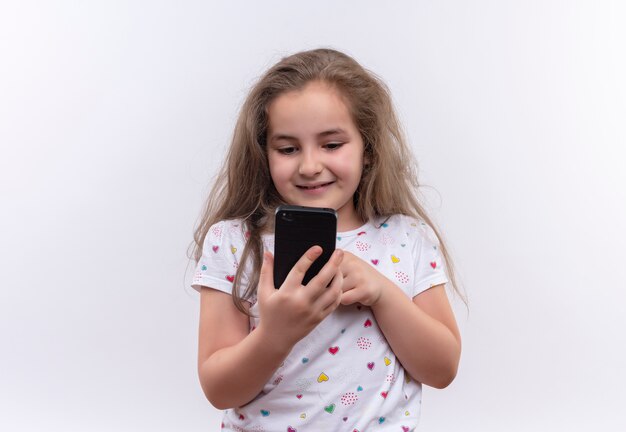 Lächelndes kleines Schulmädchen, das weißes T-Shirt trägt, das auf Telefon auf lokalisiertem weißem Hintergrund spielt