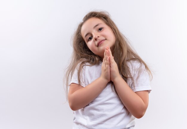 Lächelndes kleines Schulmädchen, das weißes T-Shirt trägt, das Gebetsgeste auf lokalisiertem weißem Hintergrund zeigt