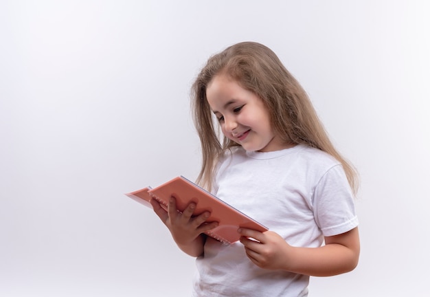 Lächelndes kleines Schulmädchen, das weißes T-Shirt trägt, das Notizbuch auf lokalisiertem weißem Hintergrund betrachtet