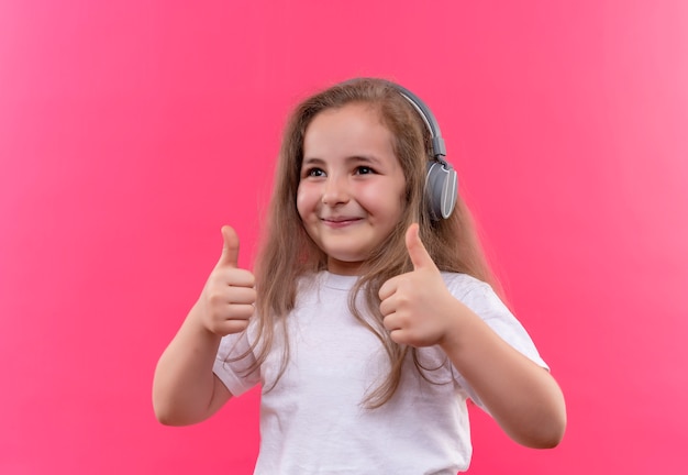 Lächelndes kleines Schulmädchen, das weißes T-Shirt trägt, hören Musik auf Kopfhörern ihre Daumen hoch auf lokalisiertem rosa Hintergrund