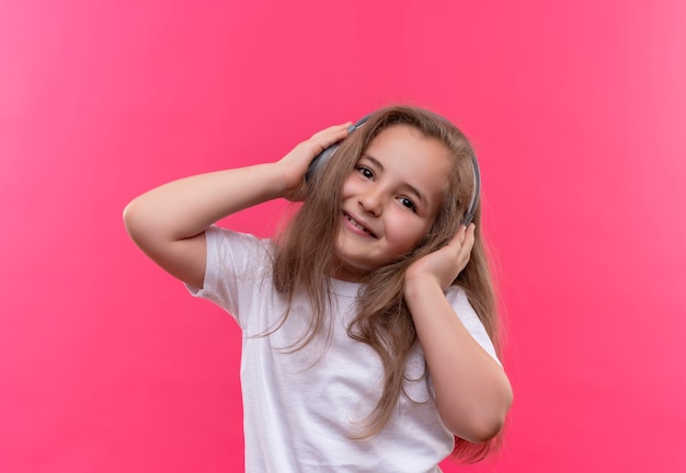 Lächelndes kleines Schulmädchen, das weißes T-Shirt trägt, hören Musik von Kopfhörern auf lokalisiertem rosa Hintergrund
