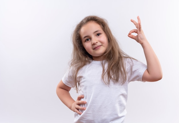 Lächelndes kleines Schulmädchen, das weißes T-Shirt trägt, legte ihre Hand auf Hüfte, die okey Geste auf lokalisiertem weißem Hintergrund zeigt