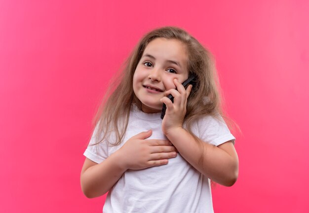 Lächelndes kleines Schulmädchen, das weißes T-Shirt trägt, spricht am Telefon auf lokalisiertem rosa Hintergrund