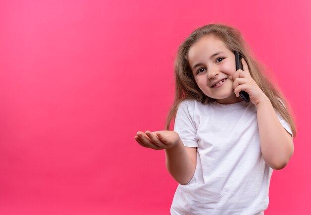 Lächelndes kleines Schulmädchen, das weißes T-Shirt trägt, spricht am Telefon gehaltene Hand, um auf lokalisiertem rosa Hintergrund weiterzuleiten