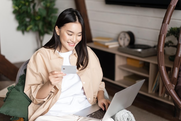 Lächelndes koreanisches Mädchen, das online von zu Hause aus auf dem Laptop kauft und eine Kreditkarte hält