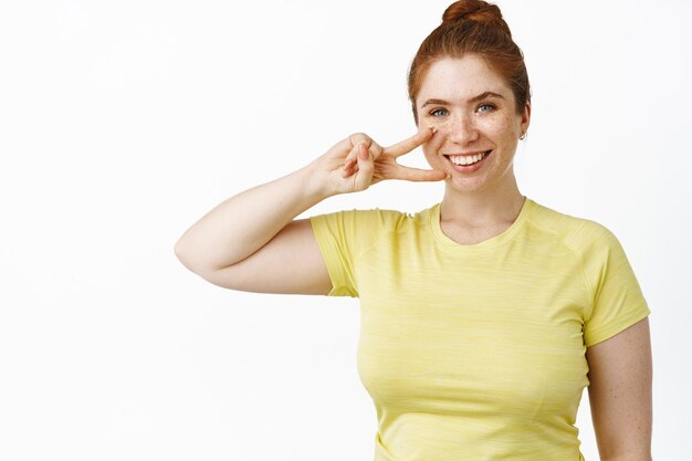 Lächelndes, kurviges rothaariges Mädchen in Sportkleidung, das Peace-Zeichen zeigt und beim Kameratraining im Fitnessstudio vor weißem Hintergrund motiviert aussieht