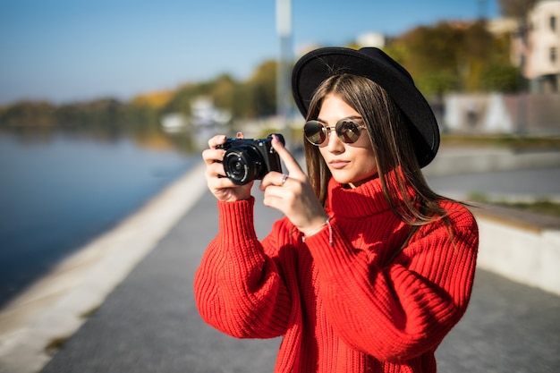Lächelndes Lifestyle-Porträt im Sommer im Freien einer hübschen jungen Frau, die sich abends in der Stadt in Europa mit einem Kamerareisefoto des Fotografen amüsiert, der Bilder in Brille und Hut im Hipster-Stil macht