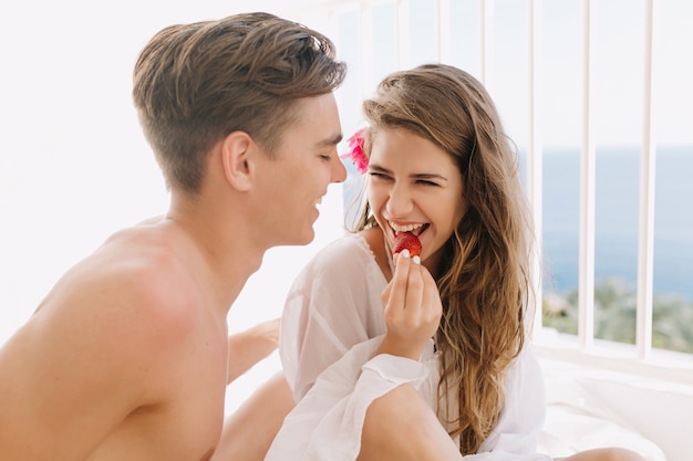 Lächelndes lockiges Mädchen mit langen Haaren, das leckere Erdbeere isst, während auf Balkon mit ihrem gebräunten Freund sitzt