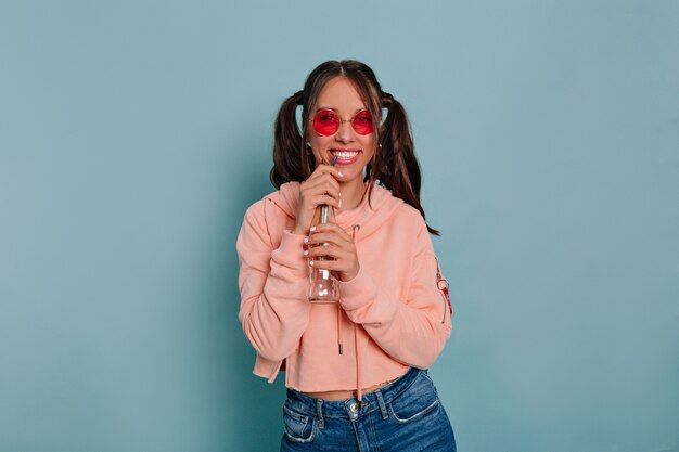 Lächelndes lustiges Mädchen mit glücklichen Gefühlen, die Zeit während des Fotoshootings im Studio genießen, das runde oink Brille und rosa Pullover trägt