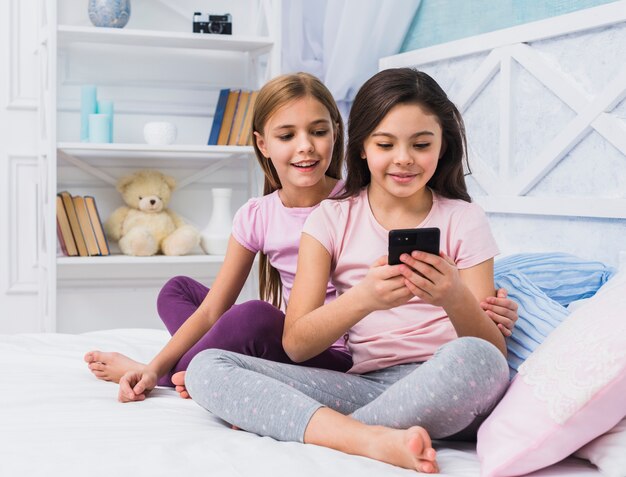 Lächelndes Mädchen, das auf dem Bett betrachtet ihren Freund unter Verwendung des Handys sitzt