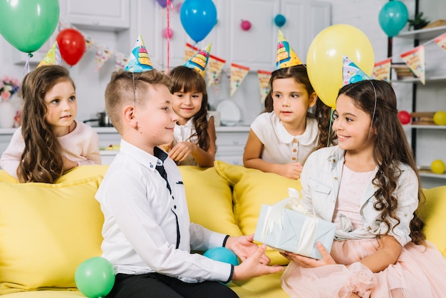 Lächelndes Mädchen, das dem Geburtstagsjungen in der Partei zu Hause Geschenk gibt