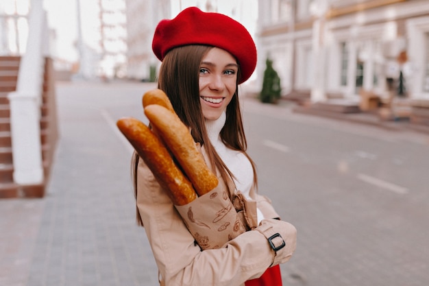 Lächelndes Mädchen, das draußen im sonnigen Morgen geht und Baguette hält.