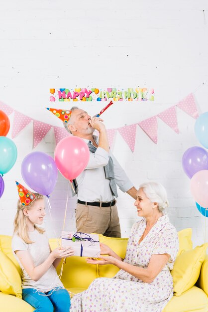 Lächelndes Mädchen, das ihrer Großmutter Geschenk vor Schlagparteihorn des Mannes gibt