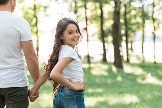 Lächelndes Mädchen, das Kamera beim Gehen in Park mit ihrem Vater betrachtet