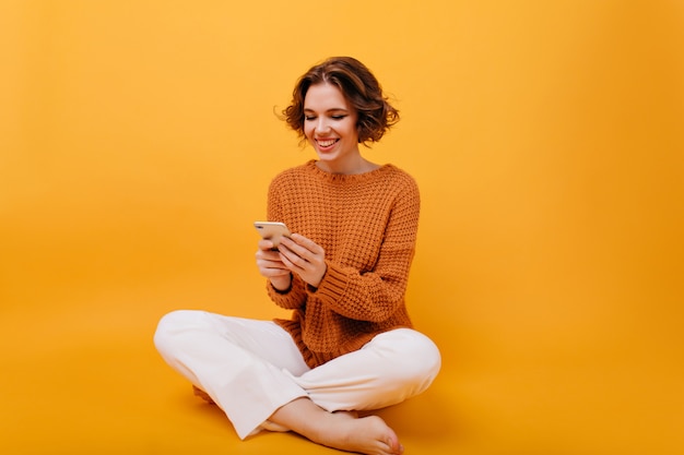 Lächelndes Mädchen im lässigen Outfit, das mit gekreuzten Beinen sitzt und Telefon hält