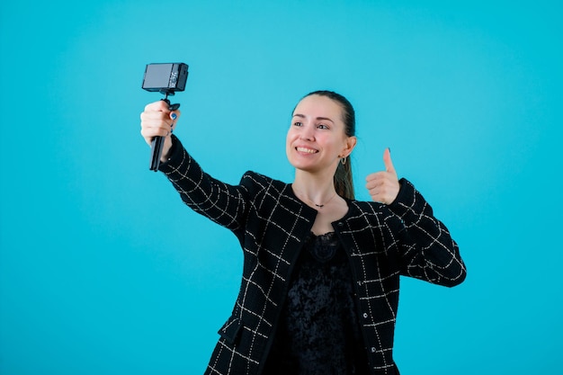 Lächelndes Mädchen macht Selfie mit ihrer Minikamera, indem es perfekte Gesten auf blauem Hintergrund zeigt