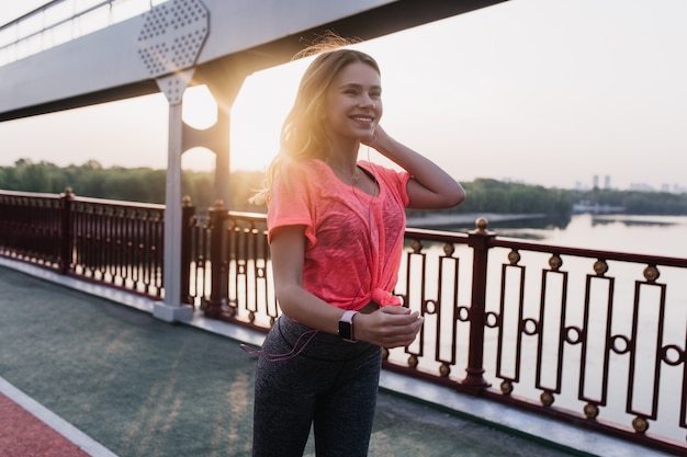 Lächelndes Mädchen mit Fitnessarmband, das im sonnigen Morgen aufwirft. Foto im Freien der freudigen jungen Dame, die Spaß nach dem Training hat.