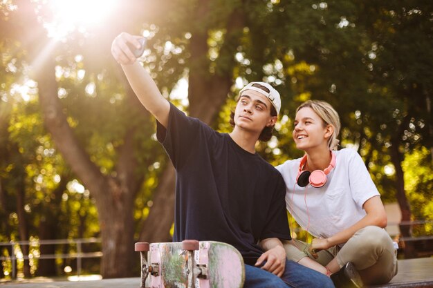 Lächelndes Mädchen mit Kopfhörern und junger Kerl mit Skateboard, der fröhlich Fotos auf der Frontalkamera des Handys zusammen im modernen Skatepark macht