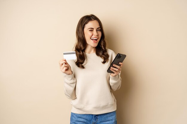 Lächelndes Mädchen mit mobiler App, Smartphone-Shopping und Kreditkarte, steht vor beigem Hintergrund, bestellt etw